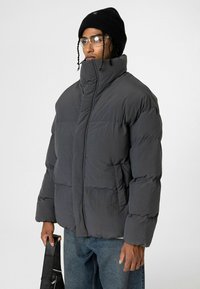 Grijze pufferjas met hoge kraag, gestikte panelen en voorzakken. Gecombineerd met blauwe spijkerbroek en een zwarte beanie, terwijl iemand een tas vasthoudt.