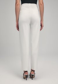 Witte op maat gemaakte broek met een gladde textuur, hoge tailleband en rechte broekspijpen, gecombineerd met zwarte gespsandalen, gezien vanaf de achterkant.