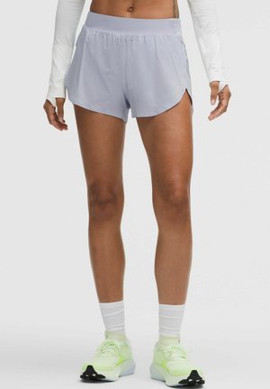 Atleta que lleva pantalones cortos de running gris claro, camiseta de manga larga blanca con agujeros para los pulgares, calcetines blancos y zapatillas de running verde neón.