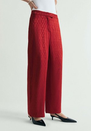 Mujer con pantalones rojos de talle alto y pernera ancha con un sutil patrón de diamantes y zapatos negros de tacón bajo con punta puntiaguda, de pie frente a un fondo sencillo.