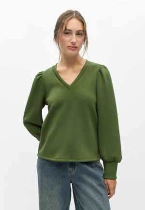 Junge Frau trägt eine grüne Bluse mit langen Ärmeln und V-Ausschnitt sowie blaue Jeans und steht vor einem schlichten weißen Hintergrund.
