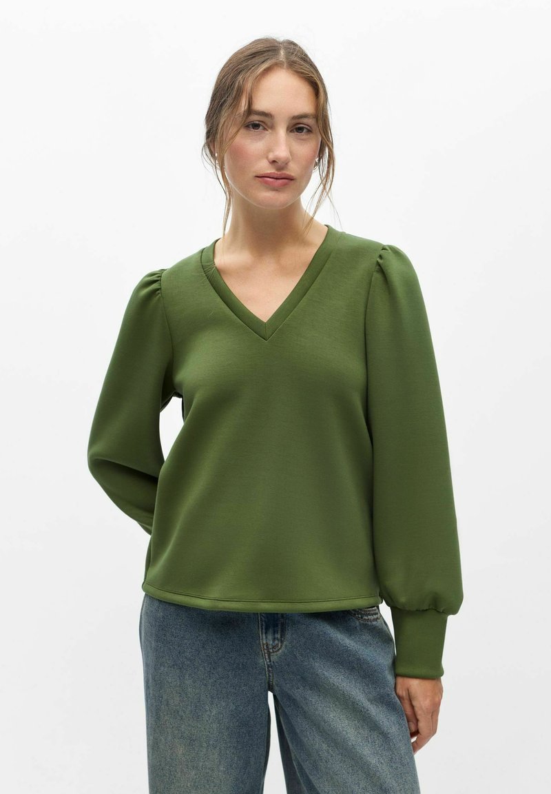 Jeune femme portant un chemisier vert à manches longues avec col en V et un jean bleu, debout devant un fond blanc uni.