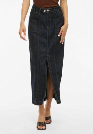 Woman wearing a dark denim midi skirt with front slit, brown sleeveless top, and brown woven open-toe heels, standing on a white background.