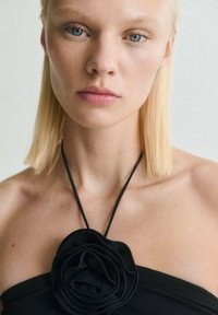Le collier floral noir présente des pétales superposés en matériau doux, attachés à un fin cordon noir, porté par un mannequin aux cheveux blonds raides.