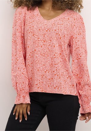 Kvinde iført orange bluse med lange ærmer med hvid paisley-mønster og sorte bukser, stående med armene afslappet ned langs siden.