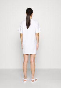 Abito t-shirt bianco in cotone con maniche corte, taglio dritto e orlo arrotondato. Indossato con sandali bianchi, capelli raccolti.