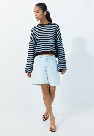 Kvinde stående med krydsede ben iført en brun og blåstribet langærmet sweater, lyse denimshorts og hvide flip-flop sandaler.