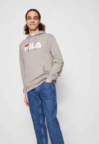 Sudadera gris con el logo "FILA" en blanco y rojo, capucha con cordones, bolsillo frontal, combinada con jeans azules, de pie contra un fondo sencillo.