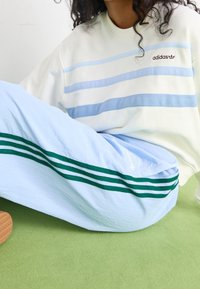 Adidas light blue pants feature green stripes, paired with a cream sweatshirt with horizontal blue stripes and a logo. Smooth texture, casual fit.