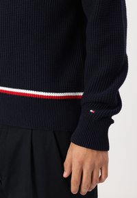 Marineblå ribbestrikket genser med hvit og rød horisontal stripe i livet og hvitt emblem på mansjetten. Strukturelt stoff.