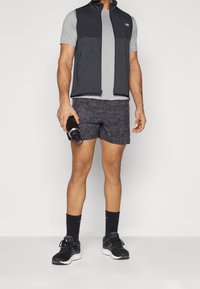 Man wearing black and gray athletic vest, gray shirt, patterned shorts, black socks, and running shoes, holding a black water bottle.