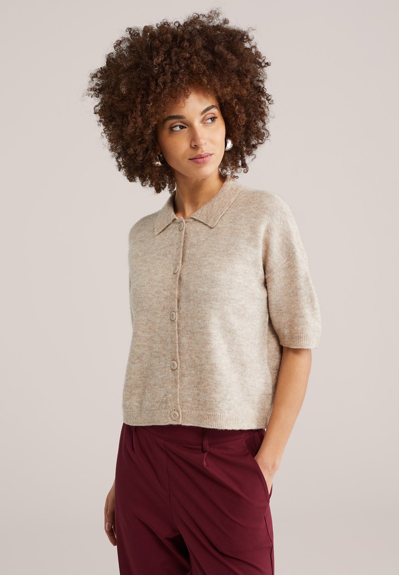 Bež kardigan kratkih rukava napravljen od mekanog, teksturiranog tkanja s kopčanjem na gumbe i klasičnim ovratnikom, nošen preko tamnoljubičastih hlača.