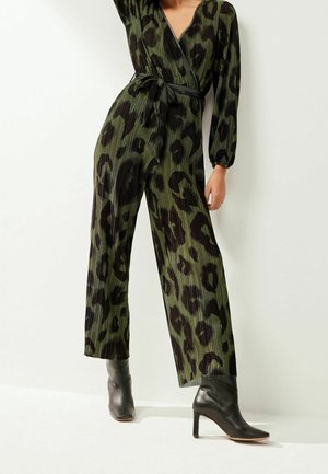 Frau trägt einen olivgrünen Wickel-Jumpsuit mit schwarzem Leopardenmuster und Falten, kombiniert mit schwarzen, hochhackigen Stiefeletten.