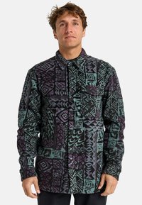 Camisa de forro polar en negro, verde azulado y púrpura con un patrón geométrico, que cuenta con un cierre de botones en el frente y un bolsillo en el lado izquierdo del pecho.