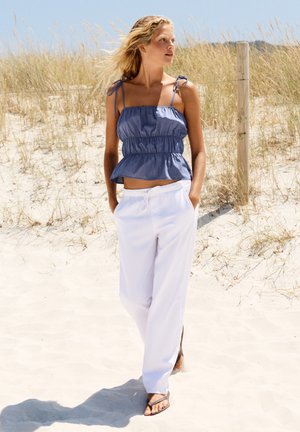 Femme aux cheveux blonds, portant un haut bleu sans manches et un pantalon blanc, marchant pieds nus sur une plage de sable avec de hautes herbes et des poteaux de clôture en bois derrière.