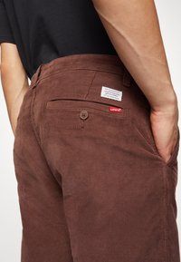 Detailní záběr osoby nosící hnědé kordové kalhoty Levi's s knoflíkem na zadní kapse a štítkem s logem, ruka zasunutá v přední kapse, viditelná černá košile.