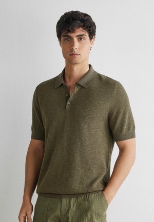 Mand med mørkt krøllet hår iført en olivengrøn kortærmet poloshirt og matchende teksturerede bukser, stående mod en ensfarvet lys væg.