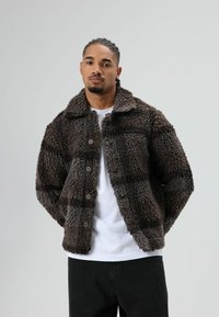 Brown faux fur jacket with a textured pattern, collared design, and button closure. Worn over a white shirt, paired with black pants.