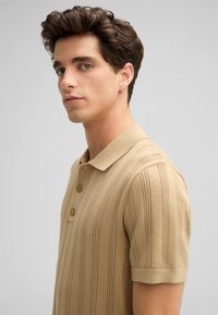 Khaki gebreide polo shirt met een gestructureerd gestreept patroon, drie knopen en een klassieke kraag. Korte mouwen met een fitted stijl.
