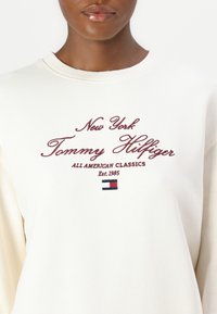 Krémová mikina s vyšitým textom "New York Tommy Hilfiger" a malým logom v červenej, bielej a modrej farbe. Mäkká textúra látky.