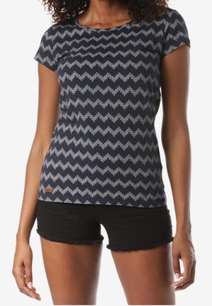 Kobieta ubrana w dopasowaną granatową koszulkę z krótkim rękawem z szarymi wzorami w zygzak oraz czarne krótkie spodenki jeansowe z postrzępionymi brzegami.