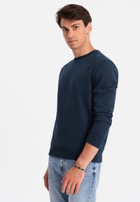 Marineblå trøje lavet af bomuldsblanding, med rund halsudskæring, ribbede manchetter og en afslappet pasform. Båret med lyseblå jeans.