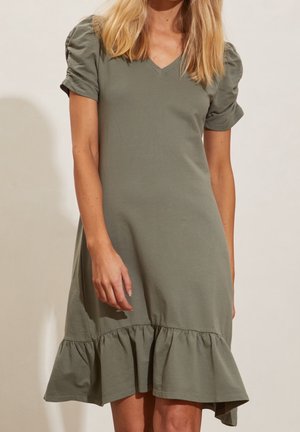 Femme portant une robe vert olive à col en V avec des manches courtes bouffantes et un ourlet volanté, debout devant un fond clair uni.