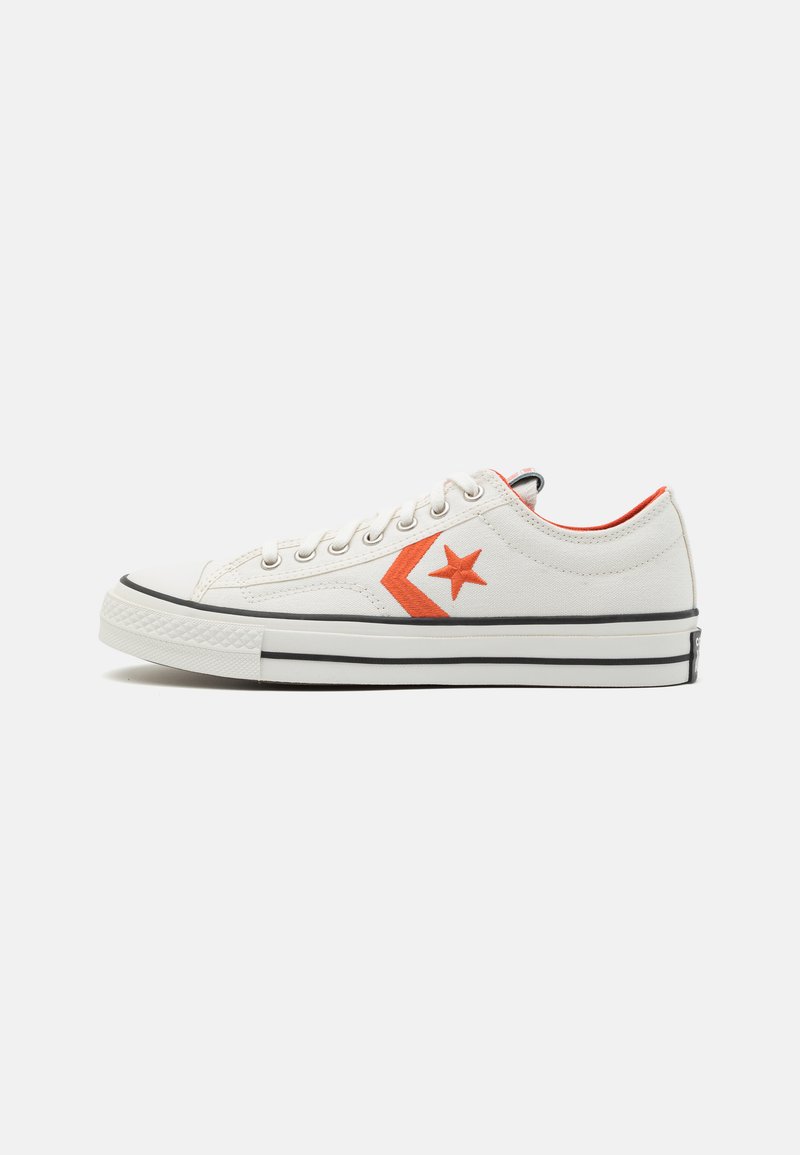 Zapatilla de lona blanca con logo de estrella naranja y detalle de chevrón, suela de goma negra y acento rojo contrastante en el collar.