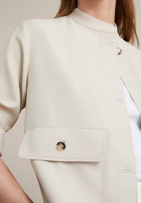 Beige kortærmet jakke lavet af glat stof, med en ståkrave, en stor klaplomme med en rund knapdetalje.