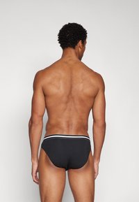 Schwarze Schwimm-Badehose mit einem weiß gestreiften Bund, in niedriger Schnittform und mit glatter Stofftextur, von hinten betrachtet.