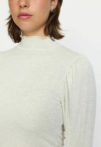 Hanorac deschis la culoare, cu mânecă lungă, gât fals, material texturat și detalii plisate la umeri. Cercei cu model argintiu vizibili.