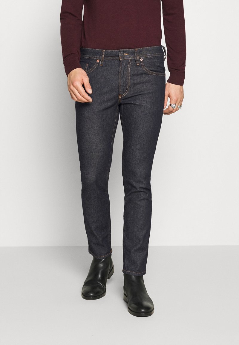 Mörk tvättade jeans med slim fit, med kontrasterande orange sömmar, standard gylf och bälteshällor. Kombineras med svarta ankelstövlar.