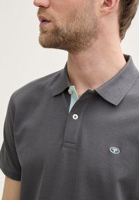 Grå polo t-shirt med struktureret stof, med en lyseblå krave og to hvide knapper. Logo på venstre brystområde.