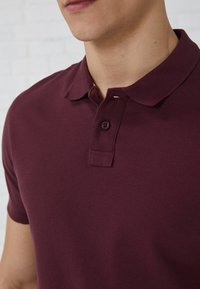 Burgundijas krāsas polo krekls, izgatavots no kokvilnas ar mīkstu struktūru, ar ribotu apkakli, divu pogu aizdari un īsām piedurknēm.