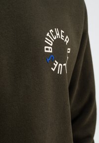 Donkergroene sweatshirt met een gestructureerde stof. Bevat een circulair logo in wit en blauw met de tekst "BUTCHER." Close-up van het linker borstgebied.