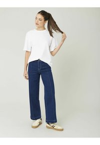 T-shirt blanc en coton à manches courtes, associé à un jean large bleu foncé avec des surpiqûres contrastées, et des baskets beiges avec des accents noirs.