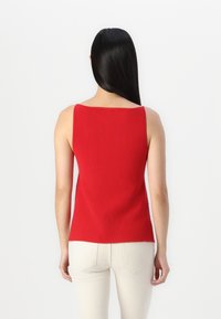Débardeur rouge à côtes avec un décolleté carré et des bretelles larges, présentant une silhouette ajustée. Porté avec un pantalon fitted de couleur claire.