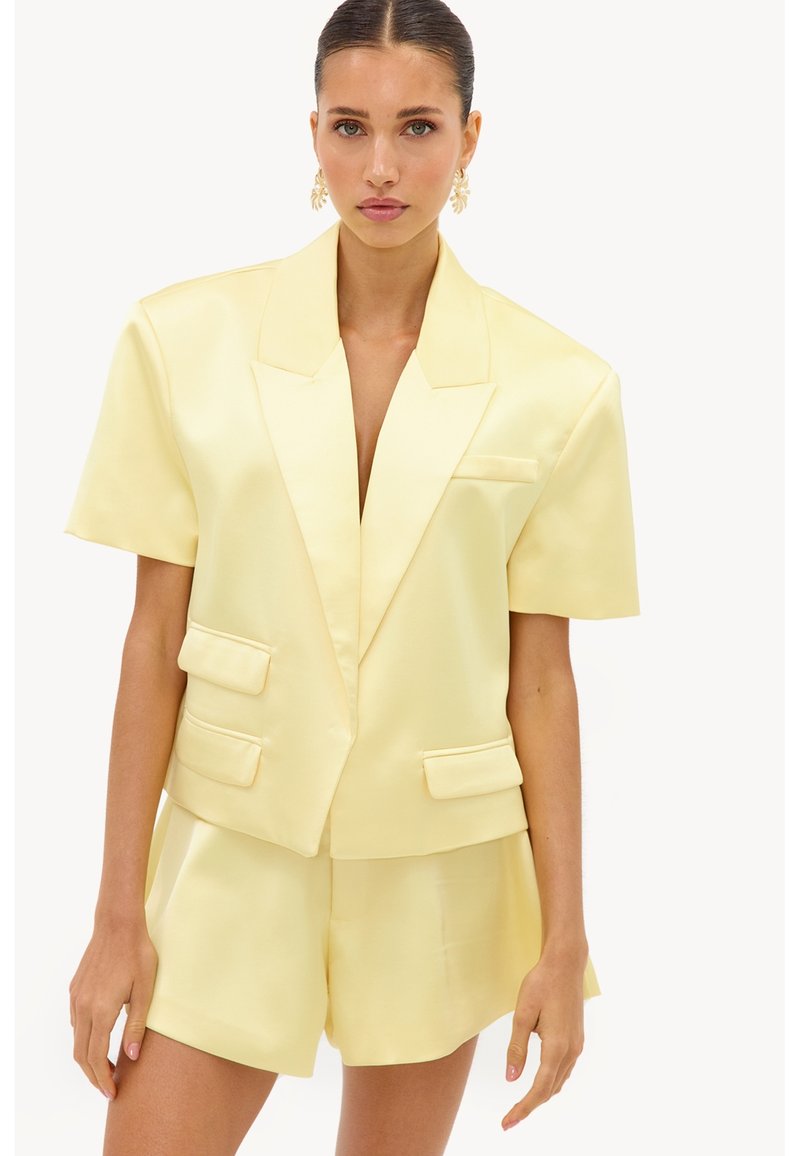 Femme portant un blazer jaune pâle à manches courtes avec un short taille haute assorti et des boucles d’oreilles dorées en forme de feuille sur fond blanc.