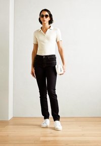 Vit ribbad polo, svarta smala jeans och vita sneakers. Accessoar: liten vit väska. Person med mörka solglasögon, inomhusmiljö.