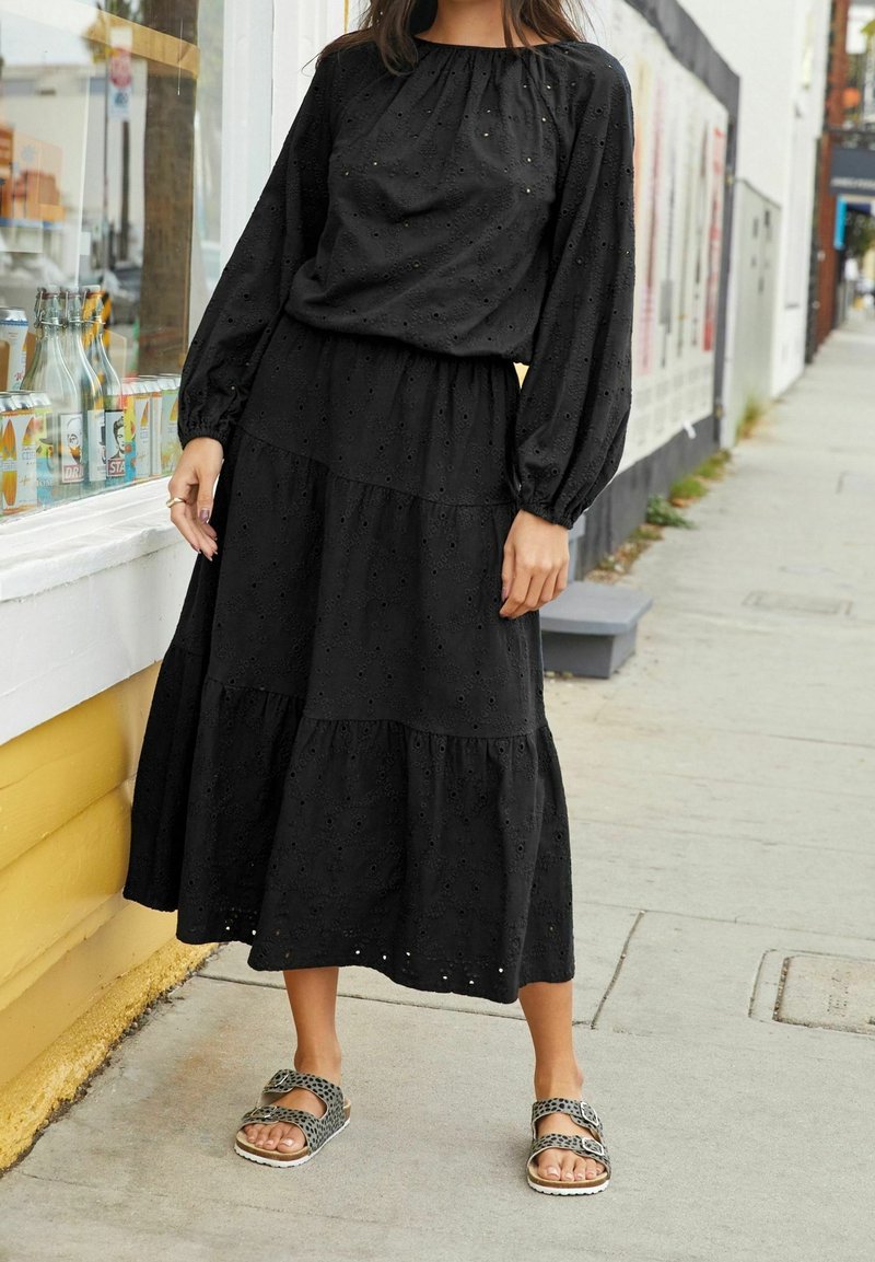 Robe noire en œillet avec manches longues, jupe à étages et encolure froncée ; assortie à des sandales noires et argentées à motif.