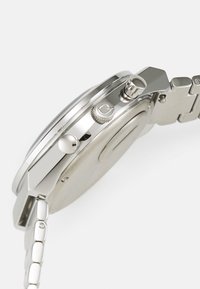 Silberner Metalluhren mit rundem Gehäuse, strukturiertem Kronen und Gliederarmband. Das Design zeichnet sich durch eine polierte Oberfläche und einen minimalistischen Stil aus.