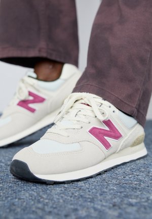 Pieds portant des baskets beiges et blanches avec un logo "N" rose sur un tapis gris, associés à un pantalon marron foncé.