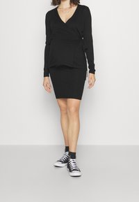 Svart stickad wrap-topp med V-urringning och långa ärmar, kombinerad med en figurnära svart kjol och svarta högkängor med vita sulor.