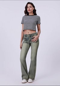 Vrouw staat in een zwart-wit gestreept crop top, vervaagde groene flared jeans en witte sneakers, tegen een effen lichte achtergrond.