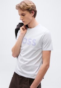 Ανοιχτό γκρι κοντομάνικο μπλουζάκι με μεγάλο, μοβ γράφημα κειμένου "OSS" στο κέντρο μπροστά, συνδυασμένο με σκούρο καφέ παντελόνι.