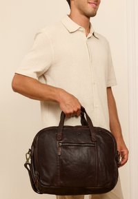 Brown leather briefcase with double handles, front zip pocket, smooth texture, rounded shape, and detachable shoulder strap.