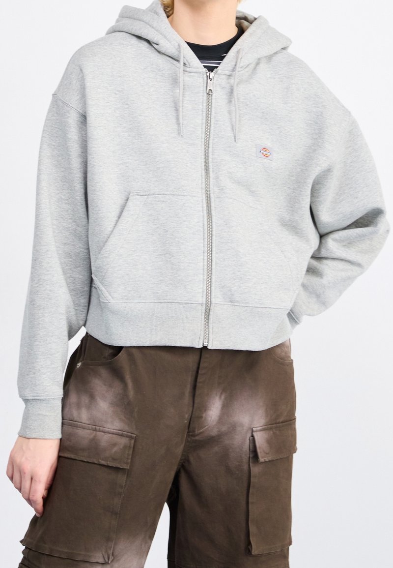 Personne portant un sweat à capuche gris clair zippé avec un petit logo et un pantalon cargo marron foncé avec de grandes poches avant.