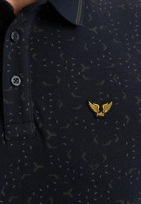 Polo navy de tejido texturizado, con un patrón sutil y un logo dorado con alas en el lado izquierdo del pecho. Los botones negros adornan el cuello.