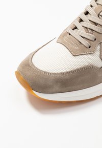 Lähikuva vaaleanbeige ja valkoisista lenkkareista, joissa on verkkokangas, mokkajäljitelmä ja vaaleanruskeat nauhat valkoista taustaa vasten.