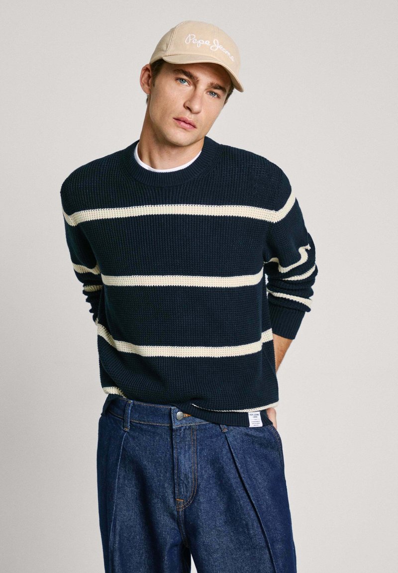 Maglione navy a maglia con righe orizzontali bianche, colletto rotondo e polsini a costine, abbinato a jeans in denim scuro a vita alta.